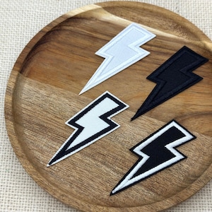 May include: Four lightning bolt patches arranged on a wooden surface. The set includes one solid white patch, one solid black patch, one white patch with a thick black border, and one black patch with a thick white border. Each patch features a stitched texture.