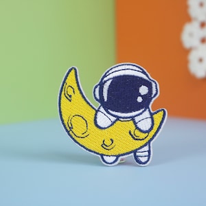 May include: An embroidered patch of a cartoon astronaut in a blue spacesuit riding a yellow crescent moon.
