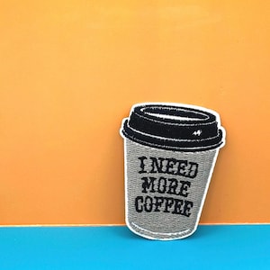 May include: A grey embroidered patch of a coffee cup with the text "I NEED MORE COFFEE" in black.