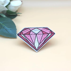 May include: Pink and white embroidered diamond shaped patch. The diamond is outlined in black thread.
