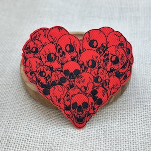 May include: A heart-shaped patch featuring a cluster of red skulls with black outlines. The patch is placed on a wooden coaster, set against a neutral fabric background. The skulls are detailed with eye sockets, noses, and teeth.