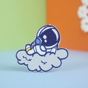 Może przedstawiać: Naszywka haftowana przedstawiająca kreskówkowego astronautę w niebieskim skafandrze kosmicznym siedzącego na białej chmurze. Astronauta ma na sobie niebieski hełm z białym wizjerem. Chmura ma niebieski kontur i białe wnętrze.