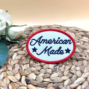 K&ouml;nnte beinhalten: Ein ovaler, bestickter Aufn&auml;her mit wei&szlig;em Hintergrund und rotem Rand. Der Text "American Made" ist in dunkelblauer Schreibschrift gestickt und wird von zwei dunkelblauen f&uuml;nfzackigen Sternen links und rechts vom Wort Made flankiert.