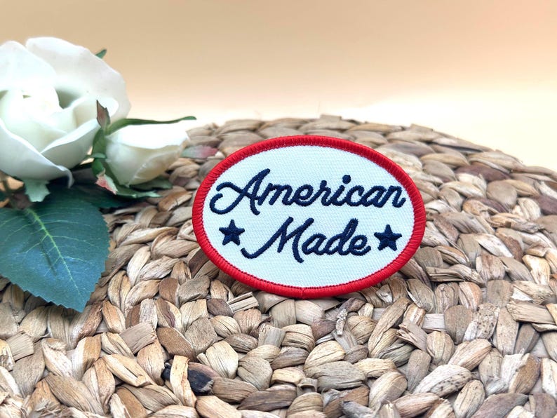 K&ouml;nnte beinhalten: Ein ovaler, bestickter Aufn&auml;her mit wei&szlig;em Hintergrund und rotem Rand. Der Text "American Made" ist in dunkelblauer Schreibschrift gestickt und wird von zwei dunkelblauen f&uuml;nfzackigen Sternen links und rechts vom Wort Made flankiert.