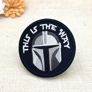 May include: A circular embroidered patch featuring a black background with a grey and white helmet design in the center. The text "THIS IS THE WAY" is stitched in white capital letters along the upper curved border of the patch.