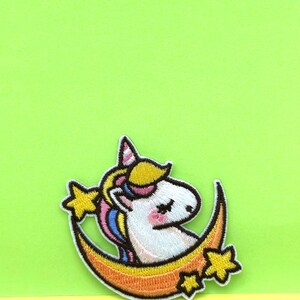 May include: A white unicorn with a rainbow mane and tail is sitting on a crescent moon with yellow stars. The unicorn has a pink nose and closed eyes.