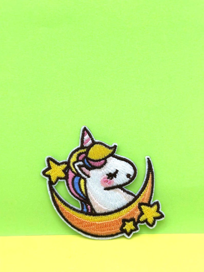 May include: A white unicorn with a rainbow mane and tail is sitting on a crescent moon with yellow stars. The unicorn has a pink nose and closed eyes.