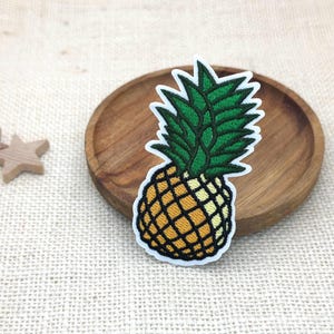 May include: An embroidered iron-on patch shaped like a pineapple. The pineapple features a yellow and orange textured body with a cross-hatched pattern and a vibrant green leafy top. The entire design is outlined in a thick white border.