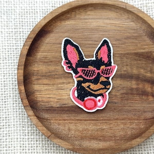 May include: Embroidered patch featuring a dog wearing pink sunglasses and headphones. The dog has black fur with pink and tan accents. The patch is on a wooden tray.