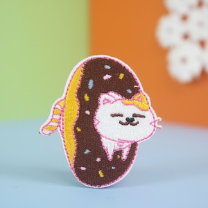 May include: Embroidered patch of a white cat with yellow ears and pink nose inside a chocolate donut with colorful sprinkles.