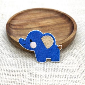 Könnte beinhalten: Ein blauer, gestickter Elefantenaufnäher mit weißem Rand, beigem Ohr und rosa Wange. Der Aufnäher liegt auf einem Holztablett mit hellbraunem Finish. Der Hintergrund ist ein strukturiertes, cremefarbenes Gewebe.