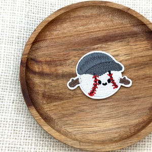 May include: A baseball-themed patch featuring a smiling baseball character wearing a gray cap. The baseball has red stitching and brown arms. The patch is on a wooden plate.