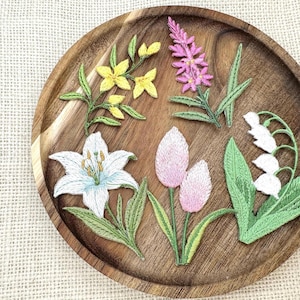 May include: Five embroidered floral patches featuring a white lily with blue accents, a pink lupine, yellow wildflowers, two light pink tulip buds, and a white lily of the valley with green leaves, all arranged on a round wooden tray.