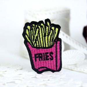 以下が含まれることがあります： ピンクの容器に入ったフライドポテトの漫画の画像が刺繍されたアイロン接着パッチ。容器には「FRIES」と印刷されています。