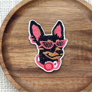May include: An embroidered patch featuring a black dog with tan accents, wearing bright pink sunglasses and a matching pink collar with a circular charm. The design is rendered in detailed thread work with vibrant pink and black color contrast.