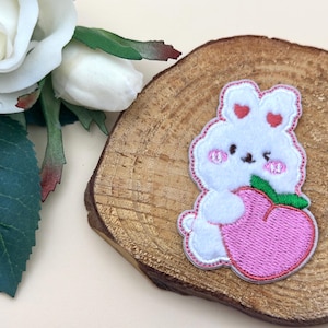 May include: A white bunny patch with pink cheeks and heart-shaped ears, holding a pink strawberry. The patch is on a wooden slice, with white roses and green leaves in the background. The patch is approximately 2 inches tall.