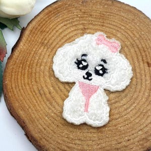 May include: Embroidered white poodle patch with a pink bow and collar. The patch features black eyes and a smiling mouth. The patch is placed on a wooden slice with a white flower and green leaves in the background.