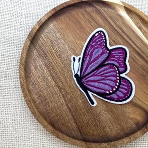 May include: A purple butterfly patch with white and black details, resting on a wooden plate. The butterfly has detailed wing patterns and a white border. The plate is made of wood.