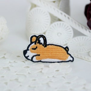 以下が含まれることがあります： 白と茶色の刺繍でできた、寝ているコーギーの犬のアップリケ。アップリケは白いレースの背景の上に置かれています。