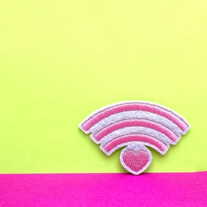 Puede incluir: Un parche bordado blanco y rosa con forma de símbolo wifi con un corazón rosa en el centro. El parche está sobre un fondo amarillo y rosa.