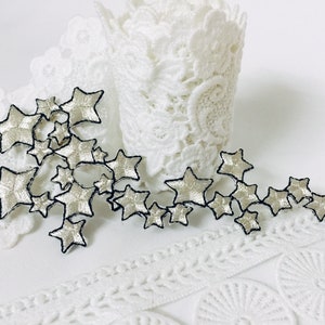 May include: A silver and black star-shaped trim with a delicate design. The trim is on a white lace background.