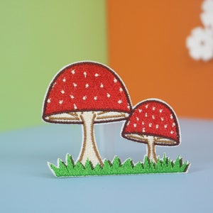 May include: Two embroidered red and white mushroom patches with brown stems and green grass. The mushrooms are on a light blue background.