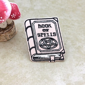 Pode incluir: Um patch rosa com um livro com as palavras "BOOK OF SPELLS" e um pentagrama. O livro tem contornos pretos e uma fita de leitura branca. No fundo, um cogumelo decorativo com chapéus vermelhos e pontos brancos.