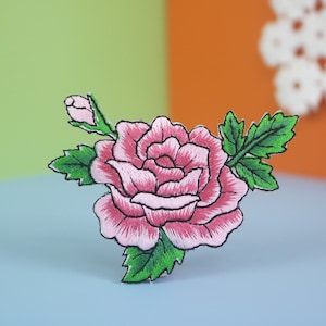 May include: Embroidered rose patch featuring a pink rose with green leaves and a bud. The patch has a black outline and is set against a colorful background. This floral patch is suitable for clothing and accessories.