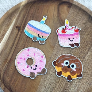 May include: Four embroidered patches featuring dessert designs. The patches include a cake with a candle, a cupcake with cherries, a pink donut with sprinkles, and a chocolate-covered pastry. Each patch has a smiling face and brown feet.