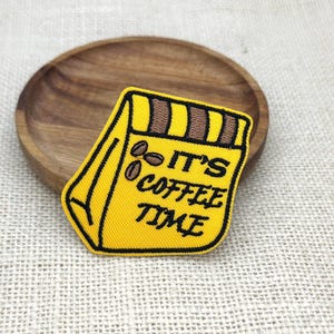 May include: A yellow embroidered patch shaped like a coffee bag with brown stripes at the top and three coffee beans. The text IT'S COFFEE TIME is stitched in black thread across the centre of the patch, which features a black border.