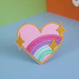 May include: A pink heart-shaped embroidered patch with a rainbow and gold glitter stars.