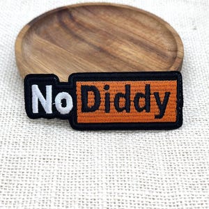 May include: A rectangular embroidered patch featuring the text No Diddy. The word No is stitched in white thread on a black background, while the word Diddy is stitched in black thread on an orange background, all enclosed by a black border.