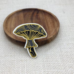 May include: An embroidered patch featuring a mushroom design with a black cap and stem accented by gold thread detailing. The mushroom cap displays intricate gold line work and a white border edge, creating a distinct contrast against the black fabric.