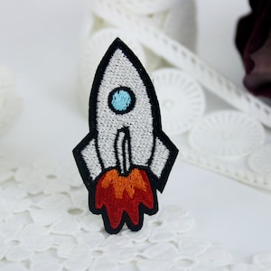 May include: White embroidered rocket patch with black outlines and orange flames. The rocket has a blue window.
