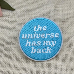 May include: A round, embroidered patch with a light blue background and white text that reads "the universe has my back." The patch has a white border and is likely for clothing or accessories.
