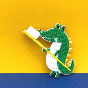 Może przedstawiać: Zielona kreskówkowa postać krokodyla trzymająca żółtą szczoteczkę do zębów z białą główką szczoteczki. Krokodyl uśmiecha się i ma biały brzuch.