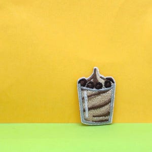 Puede incluir: Un parche bordado que representa un batido con nata montada y chispas de chocolate, en marrón y blanco.
