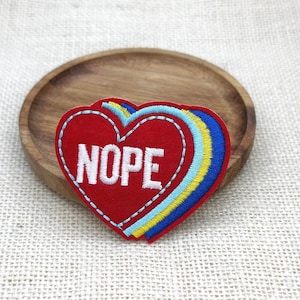 May include: A heart-shaped embroidered patch featuring a red center with the word NOPE stitched in white capital letters. The design includes layered borders in light blue, yellow, and dark blue, with a white stitched outline around the red heart.