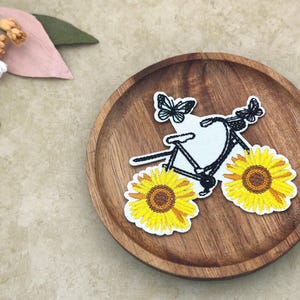 May include: Embroidered patch featuring a bicycle with sunflower wheels and butterfly accents. The bicycle is black and white, with yellow sunflowers. The patch is on a wooden plate, with a floral arrangement in the background.
