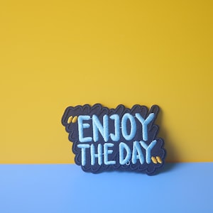 May include: A black and blue embroidered patch with the text "Enjoy the day" in a white font.