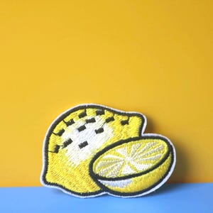 Może przedstawiać: Żółta naszywka z haftowanym cytryną i czarnym obramowaniem. Cytryna jest przekrojona na pół, pokazując wnętrze owocu.