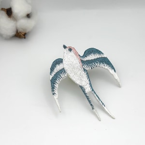 Może przedstawiać: Haftowana naszywka z jaskółką z białymi, turkusowymi i różowymi detalami. Naszywka z ptakiem ma szczegółowe szwy na skrzydłach i ciele. Jaskółka jest symbolem wolności i nadziei. Naszywkę można przyszyć lub naprasować na ubrania lub akcesoria.