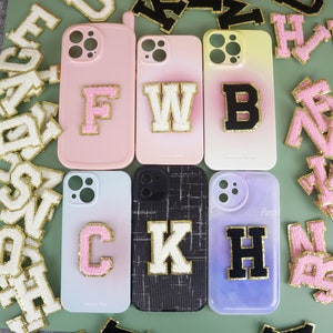 May include: Six iPhone cases with different colored backgrounds and letter patches. The letters are F, W, B, C, K, and H. The letters are in white, black, and pink with gold glitter trim.