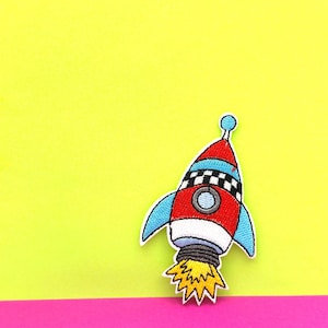 Op de afbeelding: Een rood, wit en blauw raket met een geruit patroon op de romp. De raket bevindt zich op een gele achtergrond en heeft een blauwe vlam die uit de achterkant komt.