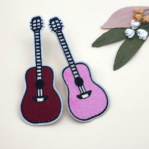 May include: Two embroidered guitar patches. One is burgundy, the other is pink, both with black and white details. The patches have a white border. The background includes decorative leaves and flowers.