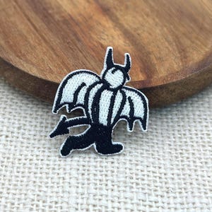 May include: An embroidered patch featuring a stylized character with white wings, a white torso, black horns, a black tail with a pointed tip, and black legs. The design is outlined in black thread, creating a high-contrast graphic appearance.
