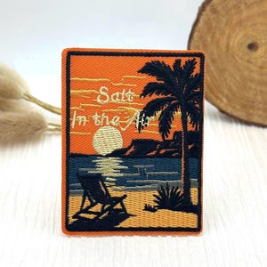Puede incluir: Parche bordado con una escena de playa. El diseño presenta una puesta de sol sobre el mar, una palmera y una silla de playa. El texto dice "Salt in the Air". El parche tiene un borde naranja.