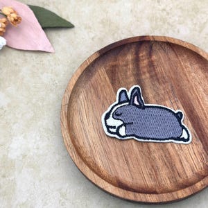 Puede incluir: Un parche bordado gris y blanco de un conejito durmiendo. El parche está sobre un plato de madera. El conejito está acostado con los ojos cerrados. El parche tiene un borde blanco.