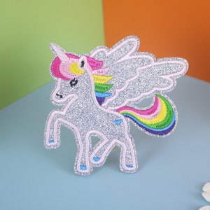 Può includere: Un unicorno di glitter argento con criniera e coda arcobaleno, e ali di glitter argento. L'unicorno è in piedi su una superficie blu chiaro.