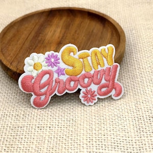 May include: An embroidered patch featuring the text Stay Groovy in yellow and pink thread. The design includes a white daisy with a yellow center, small purple star-shaped flowers, and a pink flower accent, all on a white fabric background.